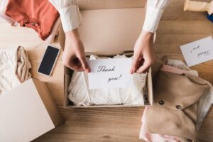 cropped view of african american owner of showroom holding card with thank you lettering near clothes, boxes and smartphone on table